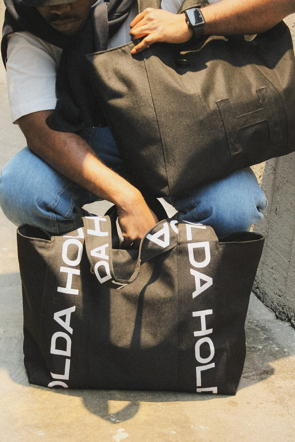 Person holding a black tote bag with 'HOLDA' branding on a concrete surface.